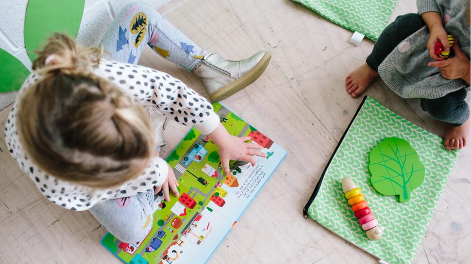 Aerial photo of a young girl reading a book on the floor and another child nearby playing with a toy with the Honey&Co Club canvas back sprawled in front with another toy on top of it