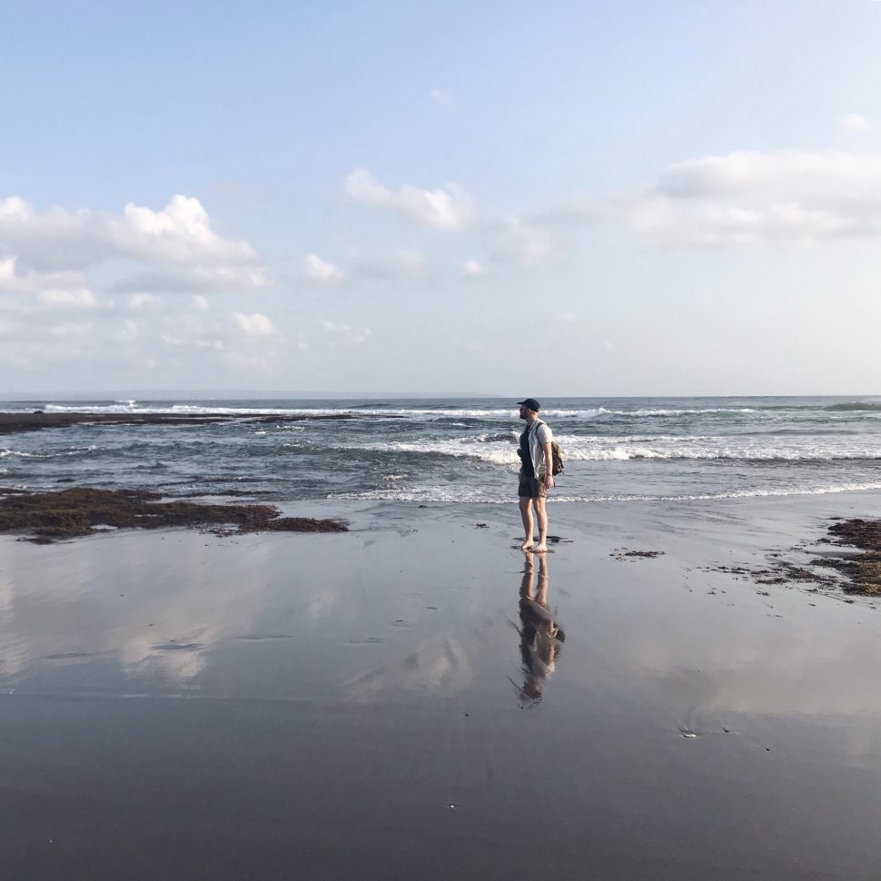 Tom stands barefoot on the sand at Canggu beach at low tide, looking out at the water. 
