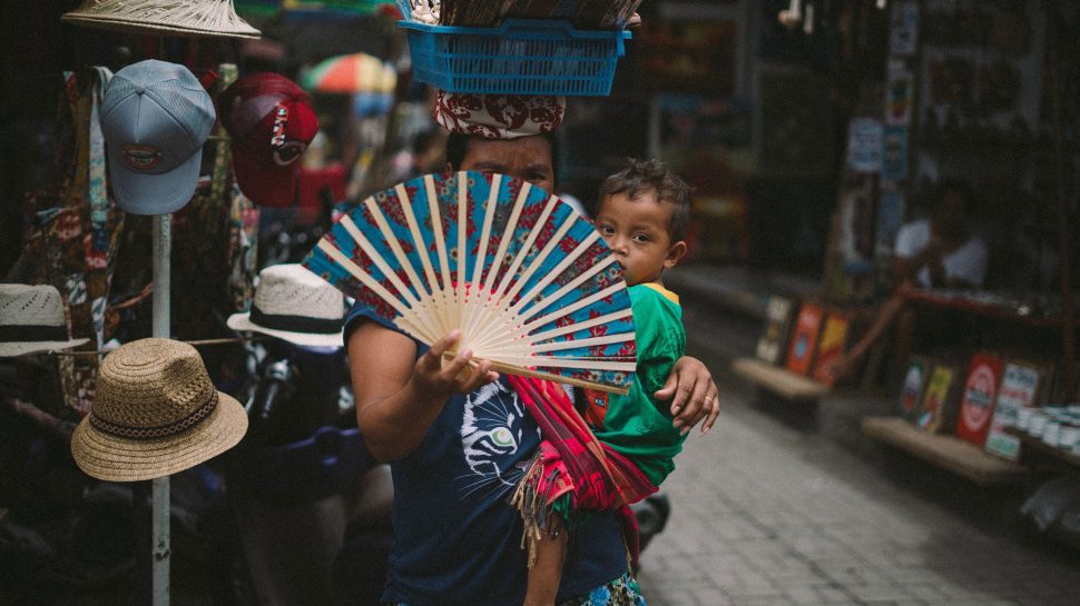 A woman in front of a hat stand in Ubud Art Market holding a fan in front of her face, balancing a basket on her head, and a young boy on her hip. 