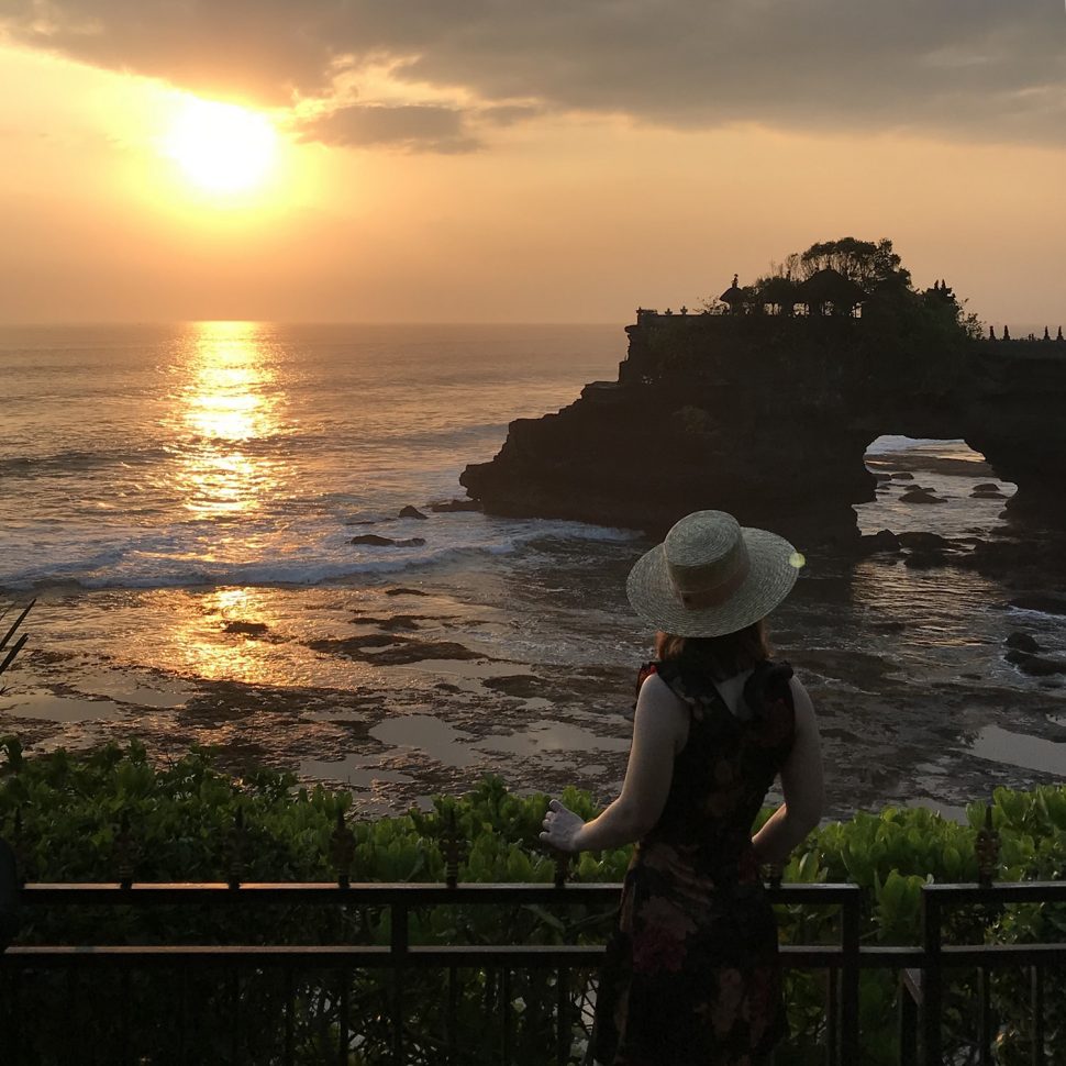 A woman looking out at the sun setting over Tanah Lot, a sacred temple in Bali. 