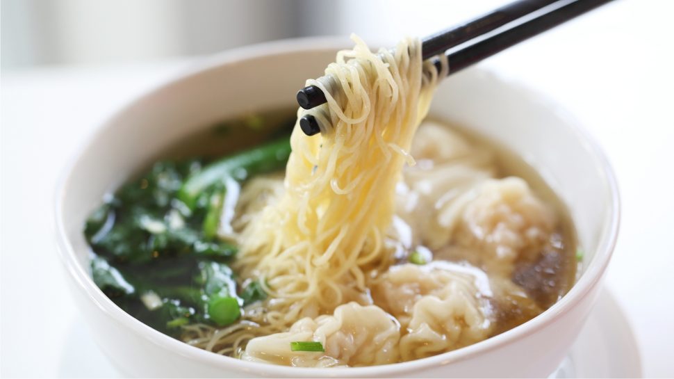 Close up of a bowl of noodles with wonton and chopsticks lifting up noodles.