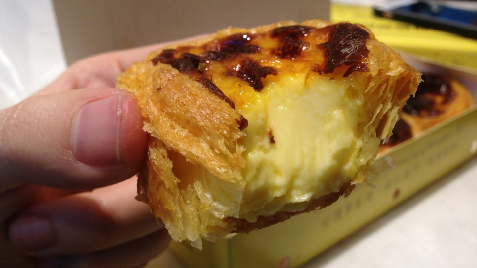 Close-up of hand holding Portuguese tart with a bite out of it showing custard filling.