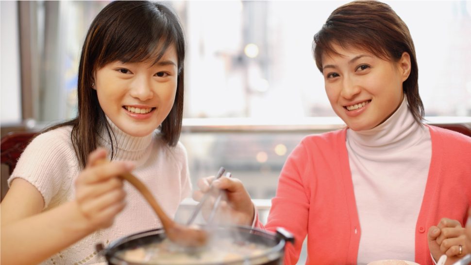 Two smiling young Chinese women stirring a pot of steaming food.