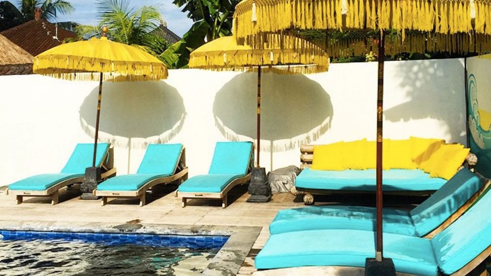 At the Chill House, bright blue deckchairs and lounges sit under yellow umbrellas around an empty pool, clear skies are in the background. 