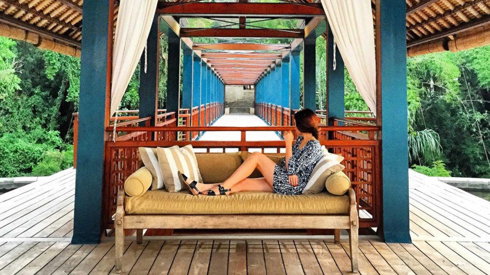 On a large canopied deck, Brooke is lounging on a couch in the foreground. The length of the deck tunnels into the distance, surrounded by forest trees. 