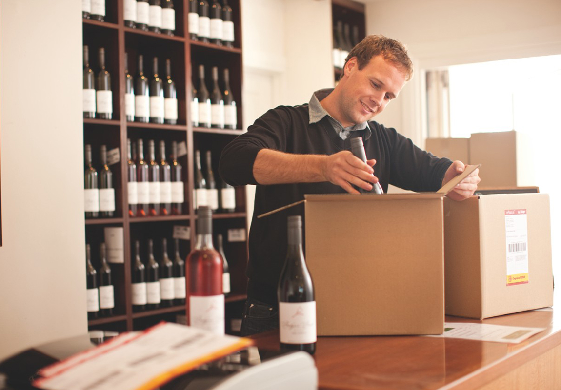 A man is standing at a counter in a wine shop and is packing wine bottles into a brown cardboard box