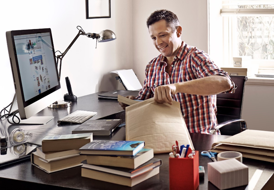 A man sitting at a desk is putting a book into a plain padded satchel