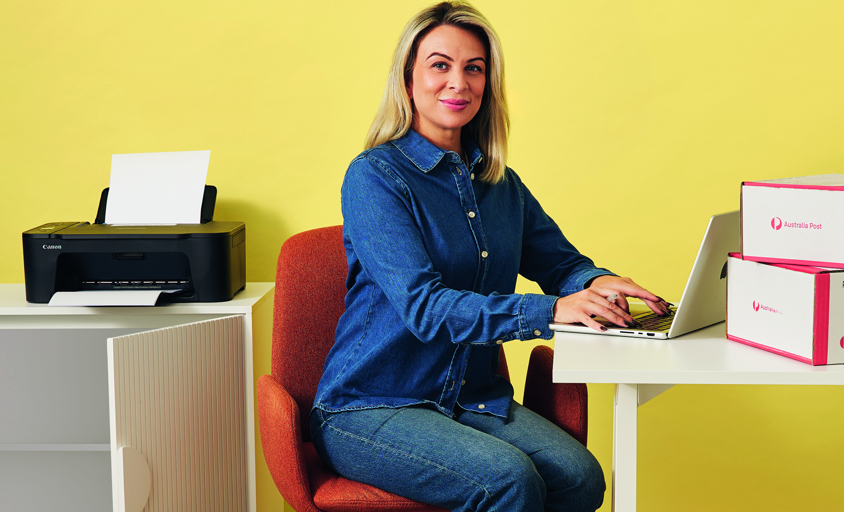Woman sitting at desk with Laptop, Post Boxes & Printer behind her.